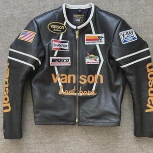 Op de afbeelding: Een zwart leren motorjack met een witte kraagrand en witte biezen. Het jack is voorzien van diverse patches, waaronder Vanson Leathers, Champion, Wiseco, K&N, Sunoco en X-Ray. De tekst Vanson Leathers is in goudkleur op de borst en mouwen gedrukt.
