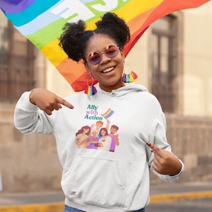 May include: White hoodie with the text "Ally with Action" and a graphic of diverse people. A rainbow flag is visible in the image, along with a rainbow flag in the background. The person in the image is wearing sunglasses and denim shorts.