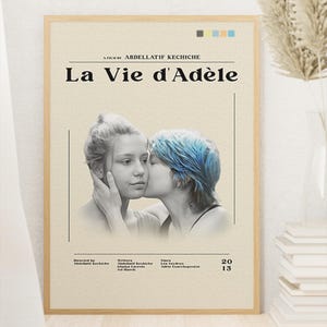 May include: A framed poster for the film "La Vie d'Adèle" featuring two women. One woman has blue hair and is kissing the other woman's cheek. The poster has text at the top and bottom, with a neutral background.