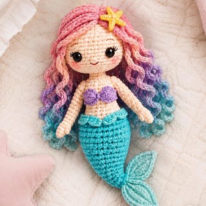May include: A crocheted mermaid doll featuring a light beige complexion, a purple shell-shaped top, and a bright turquoise tail. The doll has long, wavy hair that transitions from pink to purple to blue, accented with a small yellow starfish.