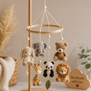 May include: A wooden mobile stand featuring six crocheted animal figures including a rabbit, elephant, bear, giraffe, panda, and lion, suspended by white strings from a circular wooden frame. A small green crocheted leaf hangs below the panda.