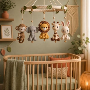 Op de afbeelding: Een hangende babymobiel met vijf gehaakte dierenfiguren: een bruine aap, een grijze olifant, een gele leeuw, een gevlekte giraf en een wit konijn. De figuren hangen aan een cirkelvormig frame dat is versierd met groene stoffen bladeren.