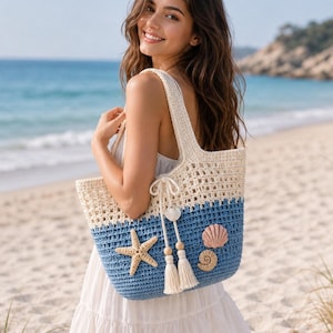Puede incluir: Un bolso tote de ganchillo bicolor con una sección superior de tejido abierto en color blanco roto y una sección inferior azul sólido. El bolso está decorado con una estrella de mar beige, una concha rosa, una caracola pequeña y dos borlas.