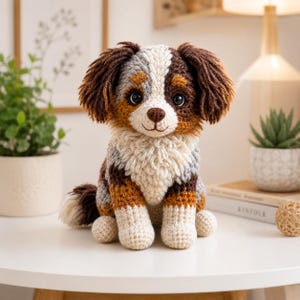 Puede incluir: Un juguete de pelaje tejido a ganchillo con forma de perro, con un pelaje tricolor de hilo marrón, blanco y gris. El animal de peluche tiene orejas largas y texturizadas de color marrón, ojos oscuros grandes, nariz pequeña y pecho blanco.