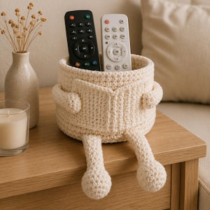May include: A cream-colored crochet basket featuring a character design with two arms holding a book-shaped front panel and two dangling legs. The basket contains one black remote control and one white remote control.