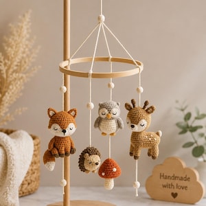 Op de afbeelding: Een houten mobielstandaard met vijf hangende gehaakte figuren: een vos, een uil, een hert, een egel en een paddenstoel. De figuren hangen aan een rond houten frame met witte koordjes en kleine houten kralen. Een houten wolkje bevat de tekst Handmade with love.