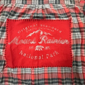 May include: A red, gray, and black plaid flannel shirt featuring a rectangular red fabric patch stitched onto the back with black thread. The patch displays white text reading Original Standard Quality Outerwear Mount Rainier National Park.