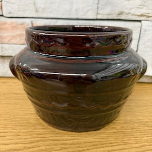 May include: A dark brown ceramic pot with a glossy finish. The pot has a wide, rounded body with decorative ridges and small handles on the sides. The rim features a wavy pattern. This vintage-style pot is suitable for kitchen or home decor.