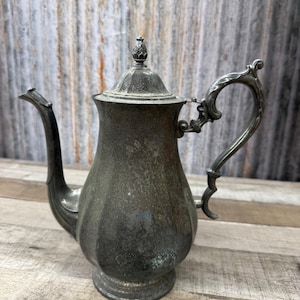 May include: A tarnished silver-toned metal teapot featuring a fluted body, a curved handle with ornate detailing, a long slender spout, and a removable lid topped with a small decorative finial. The surface shows signs of age and oxidation.