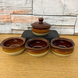 May include: A set of four ceramic bowls with a brown and tan glaze. One bowl has a lid. The bowls have a rounded shape with a handle on each side. The bowls are arranged on a wooden surface.