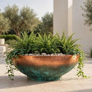 May include: A large, round, hammered metal planter bowl featuring a dual-tone finish with a polished copper upper rim and a weathered, oxidized teal-green patina on the lower section, filled with lush green ferns and small white stones.