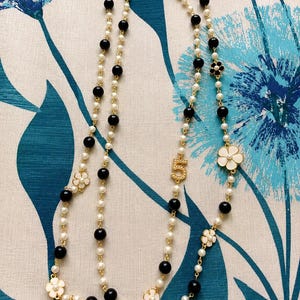 May include: A layered necklace featuring faux pearls, black beads, and gold-tone accents. The necklace includes white flower charms and a gold-tone number "5" pendant. The jewelry is displayed against a blue and white floral patterned background.