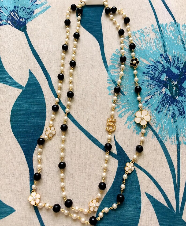 May include: A layered necklace featuring faux pearls, black beads, and gold-tone accents. The necklace includes white flower charms and a gold-tone number "5" pendant. The jewelry is displayed against a blue and white floral patterned background.