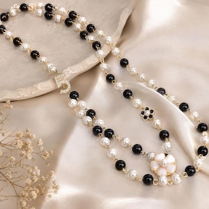 May include: A layered necklace featuring alternating black and white faux pearls, with gold-toned accents. The necklace includes a rhinestone-studded number "5" pendant and a flower-shaped pendant with white petals and a black center.