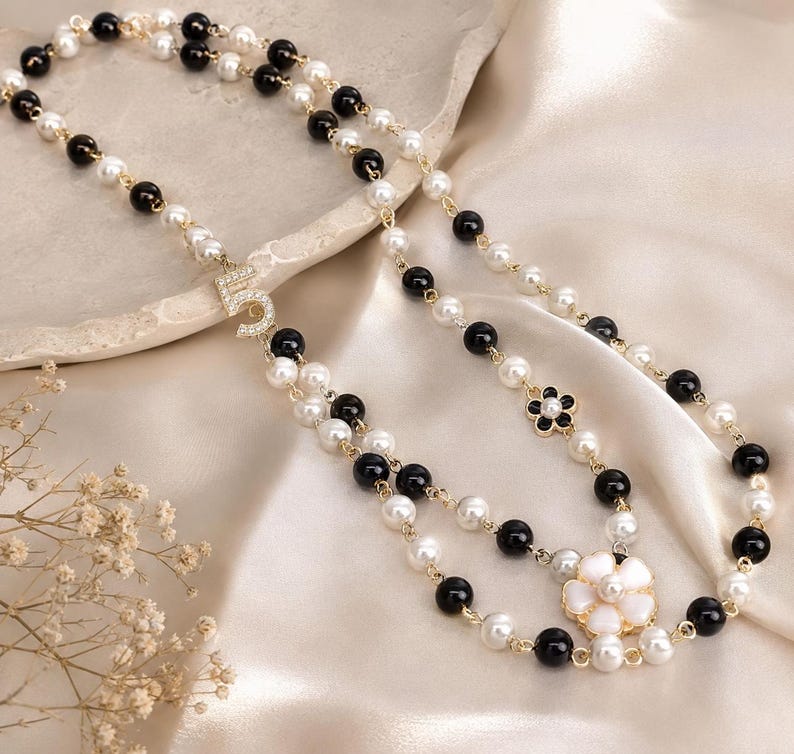 May include: A layered necklace featuring alternating black and white faux pearls, with gold-toned accents. The necklace includes a rhinestone-studded number "5" pendant and a flower-shaped pendant with white petals and a black center.