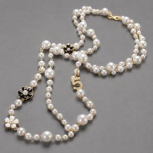 May include: A long, layered pearl necklace with gold-tone accents. The necklace features large and small white pearls, black and white flower-shaped charms, and a charm with the number "5" encrusted with small crystals. The necklace is displayed on a gray background.