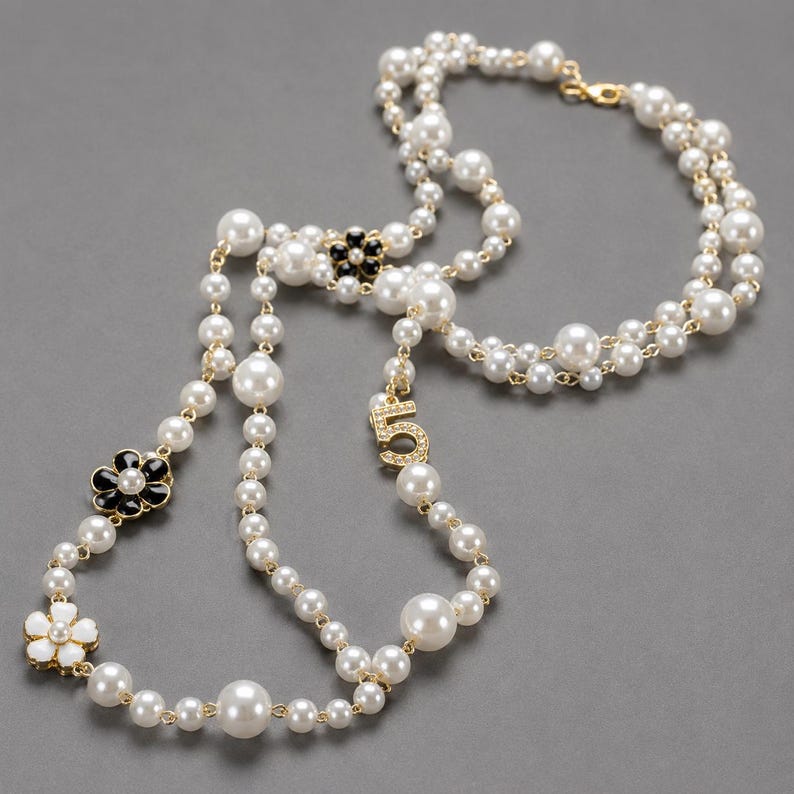 May include: A long, layered pearl necklace with gold-tone accents. The necklace features large and small white pearls, black and white flower-shaped charms, and a charm with the number "5" encrusted with small crystals. The necklace is displayed on a gray background.