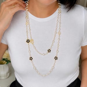 May include: A long, layered pearl necklace with gold accents and flower-shaped charms. The necklace features alternating black and white flower designs. The woman in the image is wearing a white t-shirt.
