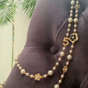 May include: A layered necklace featuring faux pearls and gold-tone accents. The necklace includes a flower and number 5 pendant. The pearls vary in size, and the necklace is draped over a dark gray surface.