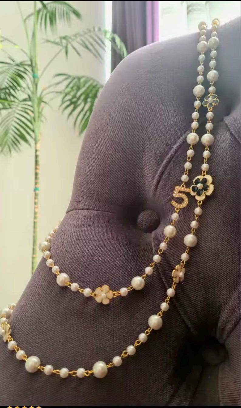 May include: A layered necklace featuring faux pearls and gold-tone accents. The necklace includes a flower and number 5 pendant. The pearls vary in size, and the necklace is draped over a dark gray surface.
