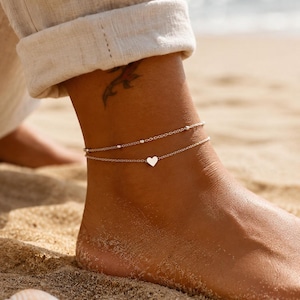 May include: Two silver anklets on an ankle at the beach. One anklet has small beads, and the other has a heart charm. The person's foot is on the sand, with a seashell in the foreground.
