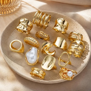 Könnte beinhalten: Eine Sammlung goldfarbener Ringe in verschiedenen Designs, darunter eine Spirale, ein Kreis und ein Ring mit einer großen weißen Perle. Die Ringe werden auf einem beigen Teller präsentiert, der ihre unterschiedlichen Formen und Texturen zeigt.