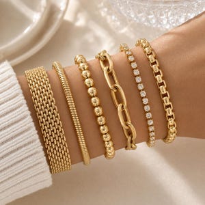 May include: An assortment of gold bracelets, featuring a mesh design, a ribbed band, beaded, chain link, a diamond-set band, and a box chain bracelet. The bracelets are shown on a wrist, highlighting their diverse textures and styles.