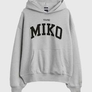 Op de afbeelding: Een lichtgrijze hoodie met een kangoeroezak aan de voorzijde, lange mouwen met geribbelde boorden en een capuchon. Op de borst staat de tekst YOUNG in kleine zwarte letters boven de grote, zwarte letters in collegestijl die MIKO vormen.