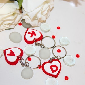 May include: Heart-shaped keychains with the letters "A", "K", and "D" in red on a white background. The keychains are red with a white heart shape. The image also includes white flowers, clear glass beads, and small red dots.