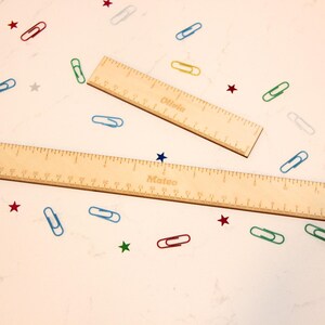 May include: Two wooden rulers with engraved measurements and names, "Olivia" and "Mateo", are displayed on a white surface. Colorful paper clips and star-shaped confetti surround the rulers, creating a playful, educational theme.