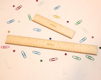May include: Two wooden rulers with engraved measurements and names, "Olivia" and "Mateo", are displayed on a white surface. Colorful paper clips and star-shaped confetti surround the rulers, creating a playful, educational theme.