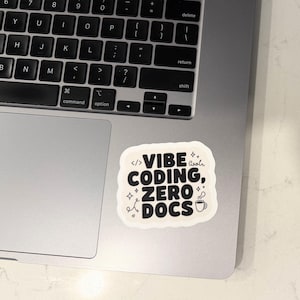 Może przedstawiać: Biała naklejka z czarnym tekstem "VIBE CODING, ZERO DOCS" umieszczona na laptopie. Naklejka ma zaokrąglony kształt i zawiera małe elementy graficzne, takie jak nawiasy kodu, gwiazdki i filiżanka kawy.