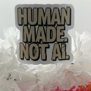 May include: A rectangular sticker with a white border featuring the beige text "HUMAN MADE. NOT AI." in a bold, blocky, distressed font with black outlines. The sticker is positioned against a background of white and pink flower petals.