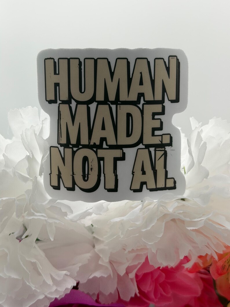 May include: A rectangular sticker with a white border featuring the beige text "HUMAN MADE. NOT AI." in a bold, blocky, distressed font with black outlines. The sticker is positioned against a background of white and pink flower petals.