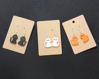 May include: Three pairs of cat-shaped earrings on display. One pair is black with green eyes, another is white, and the third is orange. Each pair is attached to a small, tan card with a silver hook.