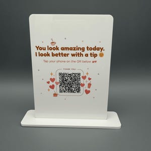 May include: A white rectangular acrylic sign with a base, featuring the brown text "You look amazing today. I look better with a tip" and "Tap your phone on the QR below". The design includes red hearts, coffee cup icons, and a central QR code.