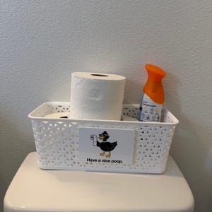 May include: A white rectangular plastic storage basket with a lattice pattern containing two rolls of white toilet paper and an orange air freshener spray. A rectangular sign on the front features a black duck wearing sunglasses and the text Have a nice poop.