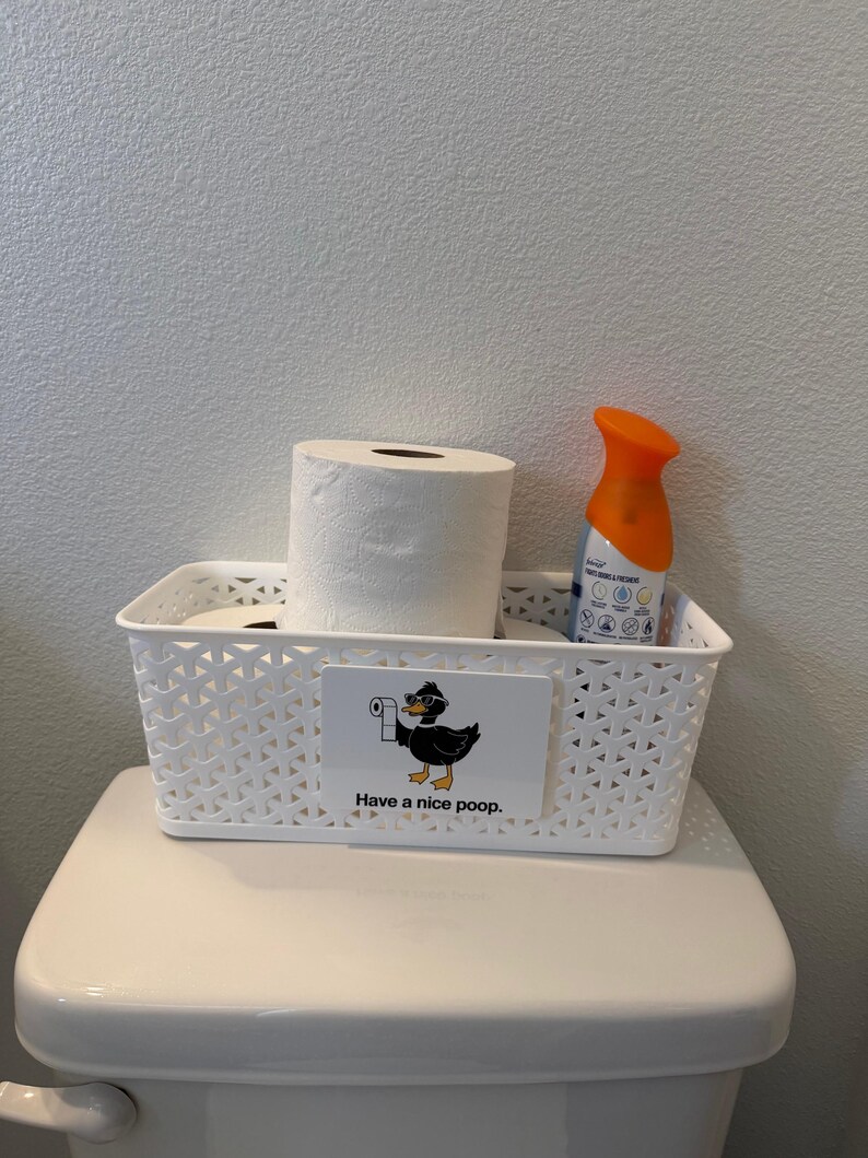 May include: A white rectangular plastic storage basket with a lattice pattern containing two rolls of white toilet paper and an orange air freshener spray. A rectangular sign on the front features a black duck wearing sunglasses and the text Have a nice poop.