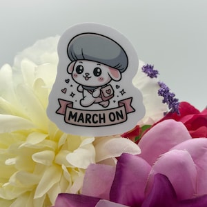 K&ouml;nnte beinhalten: Ein gestanzter Aufkleber mit einer Cartoon-Figur, die ein graues Barett und eine rosa Umh&auml;ngetasche tr&auml;gt. Unten befindet sich ein rosa Banner mit dem Text MARCH ON. Die Figur ist wei&szlig; mit gro&szlig;en schwarzen Augen, rosa Wangen und kleinen Herzen.