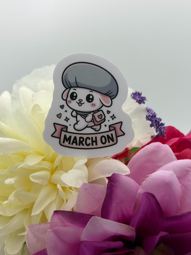 K&ouml;nnte beinhalten: Ein gestanzter Aufkleber mit einer Cartoon-Figur, die ein graues Barett und eine rosa Umh&auml;ngetasche tr&auml;gt. Unten befindet sich ein rosa Banner mit dem Text MARCH ON. Die Figur ist wei&szlig; mit gro&szlig;en schwarzen Augen, rosa Wangen und kleinen Herzen.