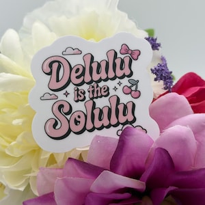 May include: A white die-cut sticker featuring the text Delulu is the Solulu in a pink and black retro-style font. The design includes small illustrations of a pink bow, two cherries, and clouds, all arranged in a playful, layered graphic layout.