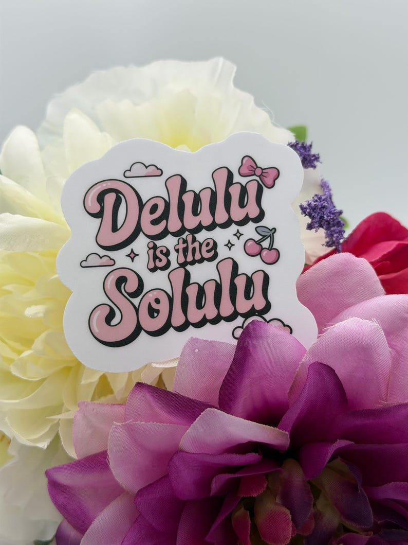 May include: A white die-cut sticker featuring the text Delulu is the Solulu in a pink and black retro-style font. The design includes small illustrations of a pink bow, two cherries, and clouds, all arranged in a playful, layered graphic layout.