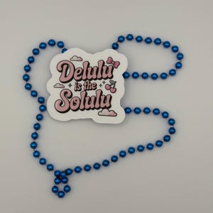 May include: A white die-cut sticker featuring the pink text Delulu is the Solulu with a small pink bow, two cherries, and cloud icons, surrounded by a long strand of shiny metallic blue spherical beads arranged in an irregular oval shape.