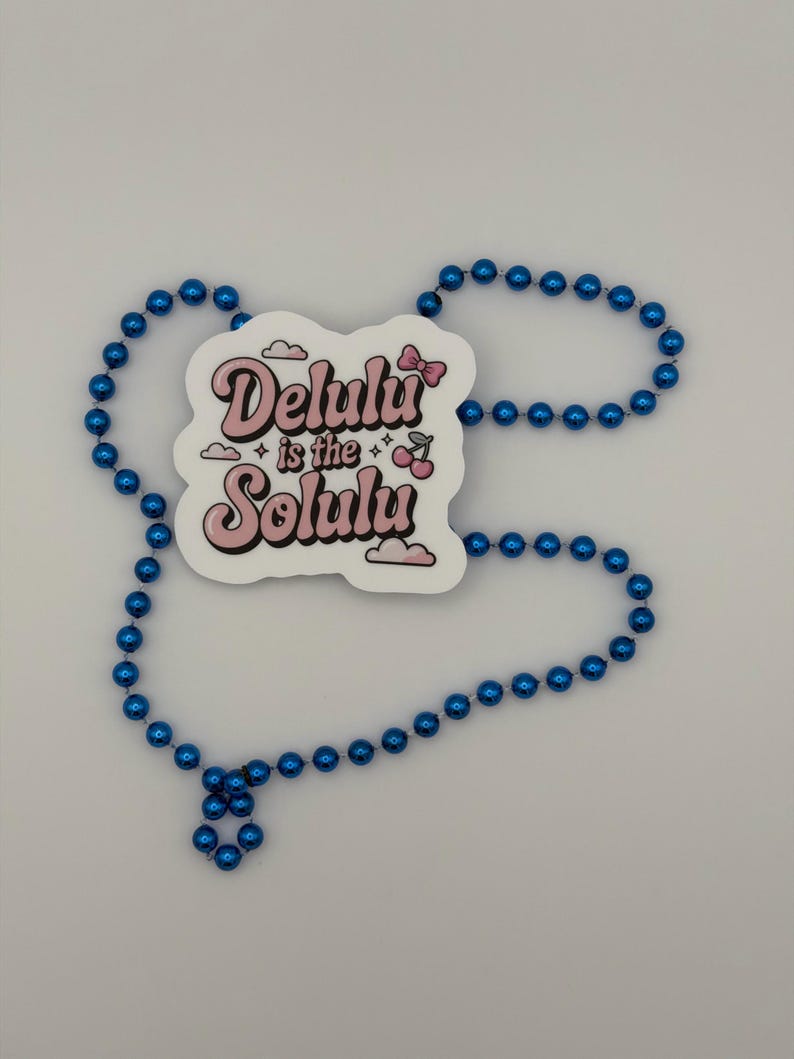 May include: A white die-cut sticker featuring the pink text Delulu is the Solulu with a small pink bow, two cherries, and cloud icons, surrounded by a long strand of shiny metallic blue spherical beads arranged in an irregular oval shape.