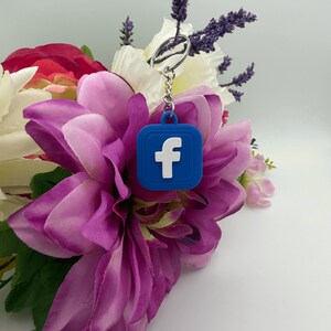 May include: A blue square keyring featuring the white letter F logo, attached to a silver metal split ring by a short chain. The keyring has a raised border design and is displayed against a background of artificial purple and white flowers.