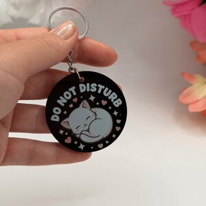 Könnte beinhalten: Ein runder schwarzer Schlüsselanhänger mit einer Illustration einer schlafenden weißen Katze in der Mitte. Der Text DO NOT DISTURB ist in weißen Großbuchstaben am oberen Rand aufgedruckt. Das Design enthält kleine rosa Herzen und Sterne.