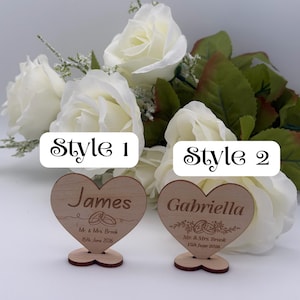May include: Two heart-shaped wooden place cards with stands. The left card features the name James, the text Mr. & Mrs. Brook, and the date 15th June 2026. The right card features the name Gabriella, the same text, and the date 15th June 2026.