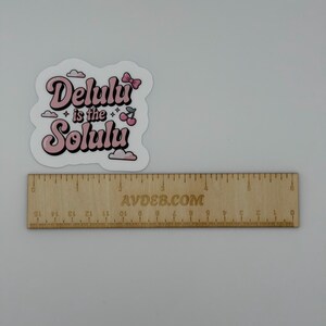 May include: A die-cut sticker featuring the pink text "Delulu is the Solulu" with a small pink bow, two cherries, and clouds. The sticker is positioned above a wooden ruler that shows the sticker is approximately 6.5 cm in width.