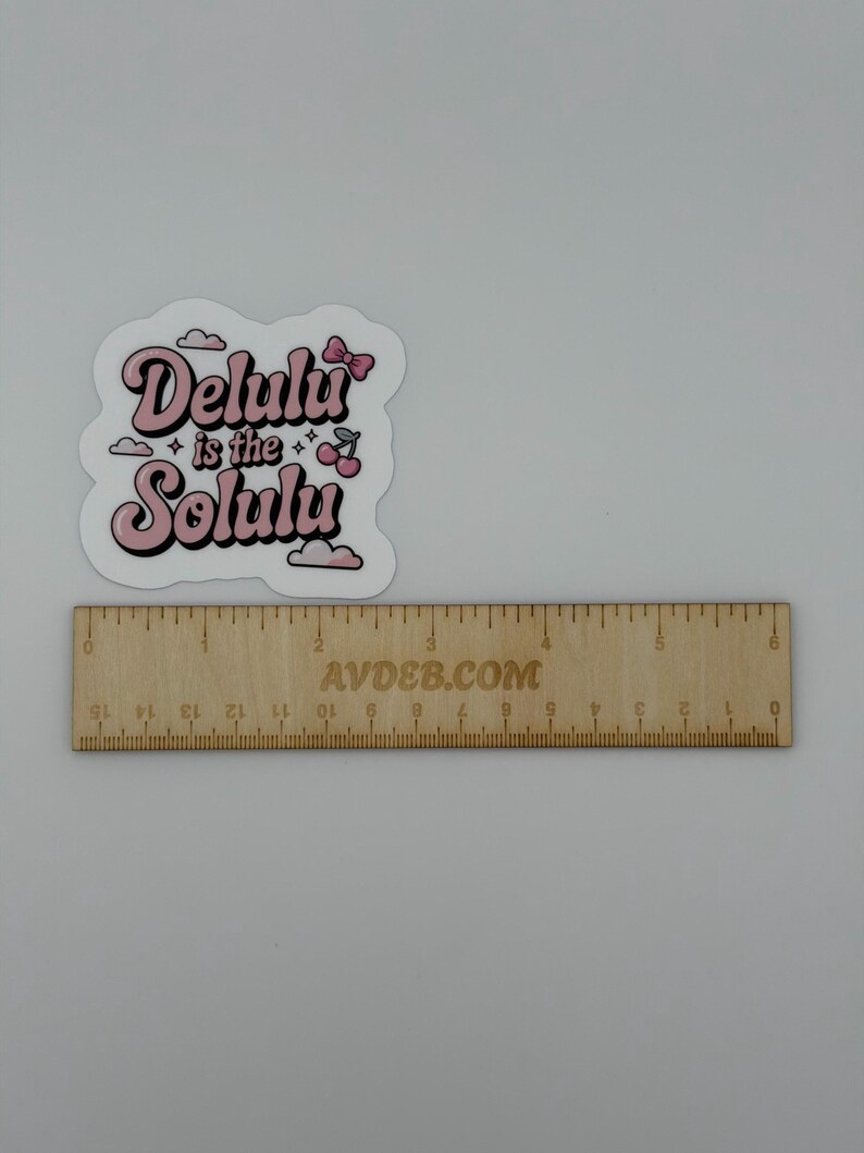 May include: A die-cut sticker featuring the pink text "Delulu is the Solulu" with a small pink bow, two cherries, and clouds. The sticker is positioned above a wooden ruler that shows the sticker is approximately 6.5 cm in width.