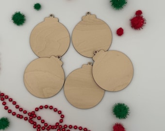 May include: Four circular wooden ornaments with small hanging loops at the top. The ornaments have a light tan, natural wood grain finish. They are arranged in a cluster, surrounded by small red and green pom-poms and a red beaded garland.